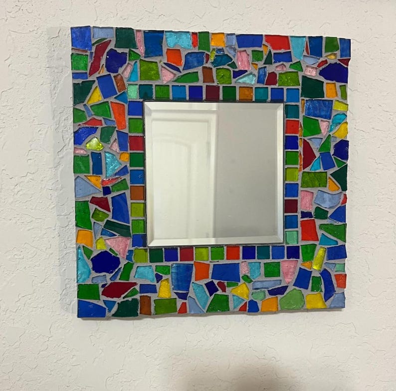 May include: A square wall mirror featuring a wide frame decorated with a colorful mosaic of irregular glass shards in shades of blue, green, red, yellow, and pink, set in gray grout, surrounding a central square reflective glass surface.