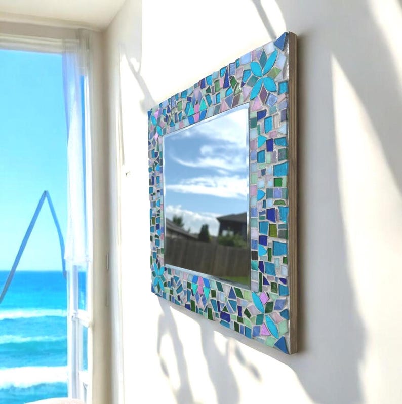 Puede incluir: Un espejo de pared rectangular con un marco de mosaico decorativo compuesto por peque&ntilde;as piezas de vidrio de formas irregulares en tonos azul, turquesa, morado y blanco transparente, dispuestas en un patr&oacute;n con motivos de p&eacute;talos.