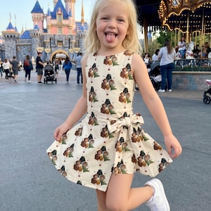 Puede incluir: Una ni&ntilde;a peque&ntilde;a con un vestido color crema con un estampado repetido de personajes de dibujos animados, posando de forma juguetona. El vestido tiene un lazo en la cintura y se combina con zapatillas blancas. El fondo incluye un castillo y un carrusel.