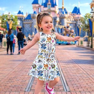 May include: A white sleeveless dress featuring colorful cartoon characters. The dress has a round neckline and a flared skirt. The girl is wearing pink sneakers. The background includes a castle and people.