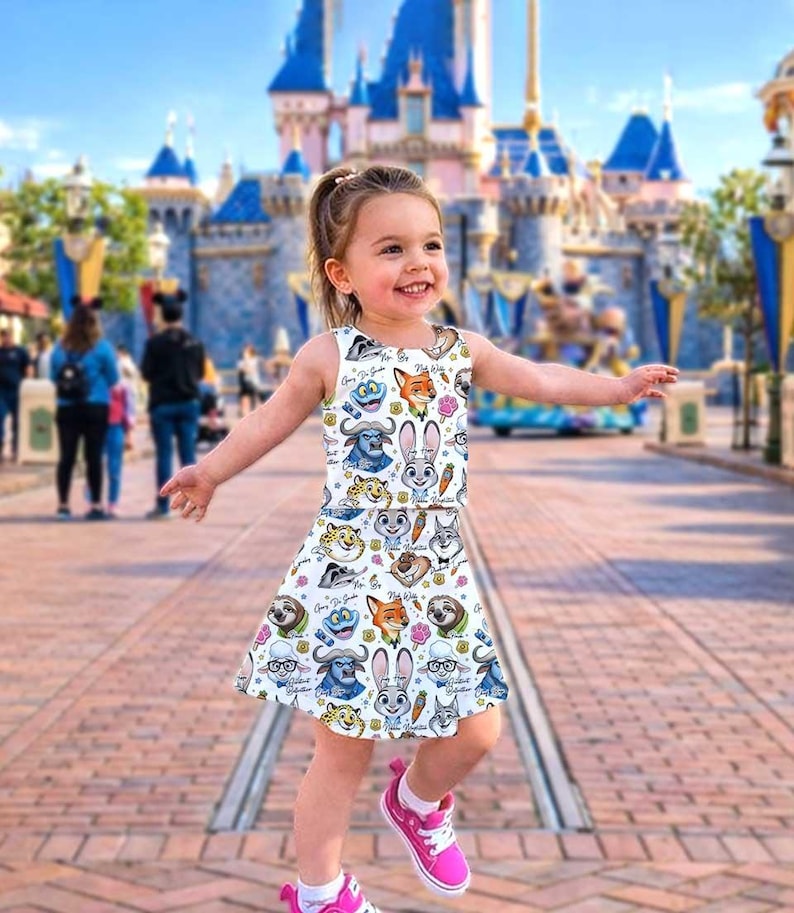 May include: A white sleeveless dress featuring colorful cartoon characters. The dress has a round neckline and a flared skirt. The girl is wearing pink sneakers. The background includes a castle and people.