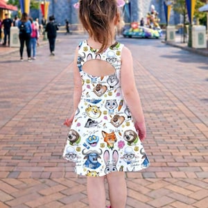 May include: A white sleeveless dress featuring cartoon characters from the movie Zootopia. The dress has a keyhole back and a flared skirt. The characters include Judy Hopps, Nick Wilde, and Flash. The dress is worn by a young child.