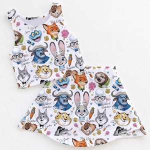 May include: White crop top and skirt set featuring a colorful pattern of cartoon animal faces. The design includes characters like a bunny, fox, and sloth, with various accents. The set is likely for children.