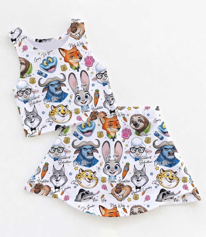 May include: White crop top and skirt set featuring a colorful pattern of cartoon animal faces. The design includes characters like a bunny, fox, and sloth, with various accents. The set is likely for children.