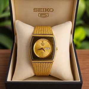 May include: A gold-tone Seiko 5 automatic watch with a square case and a textured mesh bracelet. The watch features a gold dial with baton indices, gold hands, and a day-date window at the three o'clock position, presented in a black display box.