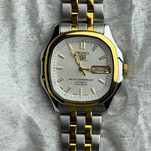 May include: A two-tone Seiko 5 Superior automatic watch with a silver-colored textured dial, gold-tone hands, and hour markers. The watch features a day-date window at the three o'clock position and a stainless steel link bracelet with gold-tone accents.