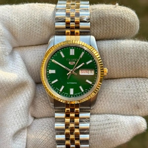 May include: A Seiko 5 automatic wristwatch featuring a vibrant green dial with silver-tone hour markers and hands. The watch has a two-tone stainless steel and gold-tone fluted bezel and a matching jubilee-style link bracelet with a metallic finish.