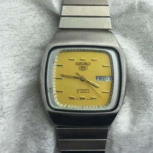 May include: A Seiko 5 automatic wristwatch with a square-shaped silver-tone stainless steel case and matching link bracelet. The watch features a bright yellow dial with gold-tone hour markers, hands, and a day-date window at the three o'clock position.