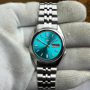 May include: A Seiko 5 automatic wristwatch featuring a vibrant turquoise sunray dial with silver-tone baton indices and hands. The watch includes a day-date window at the three o'clock position and a polished stainless steel link bracelet.