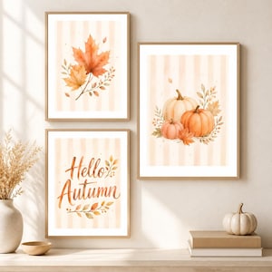 May include: Three framed wall art prints featuring watercolor illustrations of autumn themes. One print shows orange maple leaves, another displays three pumpkins with foliage, and the third features the text Hello Autumn with leaf accents.