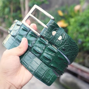 May include: A dark green belt with a textured, embossed pattern resembling reptile scales. The belt features a polished silver-tone rectangular metal buckle and two silver-tone metal rivets securing the strap near the buckle attachment point.