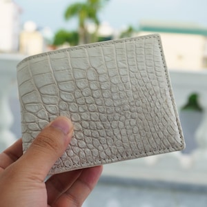 Peut inclure: Un portefeuille pliable présentant un extérieur texturé blanc cassé avec un motif gaufré distinct imitant l'apparence de la peau de crocodile. Le portefeuille présente des coutures visibles sur les bords et une forme rectangulaire.