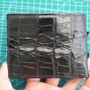 May include: A black bifold wallet featuring a glossy, textured surface with a distinct scale pattern. The exterior displays a grid-like arrangement of rectangular scales, characteristic of a reptile skin texture, with a polished finish.