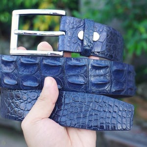 Peut inclure: Une ceinture bleu foncé présentant un motif texturé en relief rappelant le cuir exotique. La ceinture comprend une boucle en métal argenté poli avec un cadre rectangulaire et un ardillon, fixée par un passant en cuir bleu assorti.