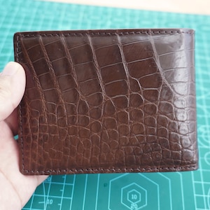 Könnte beinhalten: Eine dunkelbraune Geldbörse im Bifold-Design mit einer texturierten Krokodilleder-Optik. Das Portemonnaie hat eine rechteckige Form mit sichtbaren Nähten an den Kanten und einer glänzenden Oberfläche, die ein detailliertes Schuppenmuster zeigt.