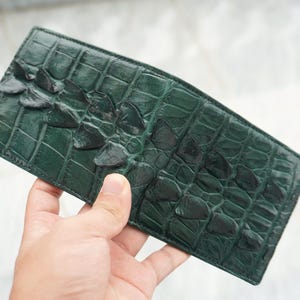 May include: A rectangular dark green wallet featuring a textured, raised scale pattern across the surface. The wallet displays a distinct row of prominent, darker green ridges running horizontally along the top section of the exterior leather.