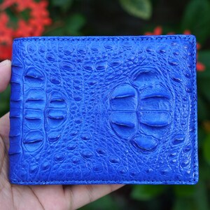 May include: A vibrant blue bifold wallet featuring a prominent, raised crocodile skin texture. The surface displays a detailed, repeating pattern of scales and ridges characteristic of exotic leather, with a consistent deep blue color throughout.