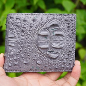 Puede incluir: Una cartera plegable de cuero gris texturizado que presenta un patrón de escamas en relieve en toda la superficie. El material muestra una textura orgánica distintiva con protuberancias y crestas de diversos tamaños.