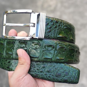 Op de afbeelding: Een donkergroene riem met een gestructureerd, reliëfpatroon dat het uiterlijk van reptielenschubben nabootst. De riem is voorzien van een gepolijste, zilverkleurige metalen gesp met een rechthoekig frame en een geribbeld detail aan de zijkant.