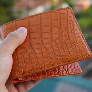 May include: A bi-fold wallet in a warm tan color featuring a distinct embossed crocodile skin texture. The wallet has visible stitching along the edges and a rectangular shape with rounded corners, showcasing a detailed scale pattern.