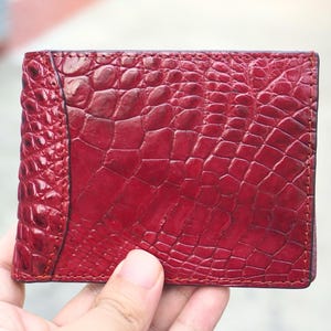 Può includere: Un portafoglio bifold rosso intenso caratterizzato da una texture in rilievo che imita la pelle di coccodrillo. Il portafoglio presenta un design a pannelli distinto sul lato sinistro con squame più piccole, che sfumano in motivi rettangolari.