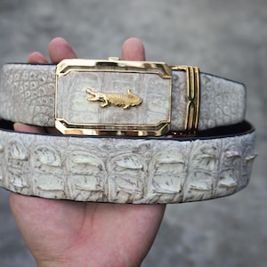 Pode incluir: Um cinto bege claro com um padrão texturizado semelhante a escamas e uma fivela retangular dourada proeminente com um pequeno emblema de crocodilo dourado em relevo no centro. A tira do cinto apresenta uma textura rígida e saliente.