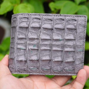 Puede incluir: Una cartera rectangular plegable en un tono gris apagado, con una superficie texturizada que presenta crestas horizontales elevadas en forma de escamas en todo el panel frontal. El material muestra un patrón segmentado con sutiles tonos verdes.