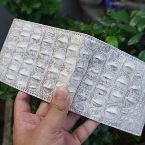 Può includere: Un portafoglio pieghevole caratterizzato da una texture in pelle di coccodrillo color bianco sporco e grigio chiaro con squame in rilievo visibili. Il portafoglio ha una forma rettangolare con bordi cuciti e una finitura in pelle di rettile.