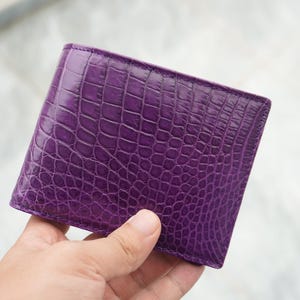 May include: A bi-fold wallet in a vibrant purple colour featuring a textured, embossed pattern that mimics the appearance of crocodile skin. The wallet has a rectangular shape with clean, stitched edges and a glossy finish on the exterior surface.