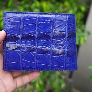 May include: A vibrant royal blue bifold wallet featuring a textured, raised scale pattern across the exterior surface. The wallet displays a distinct, repeating rectangular scale design with a glossy finish, showcasing the natural grain and texture.