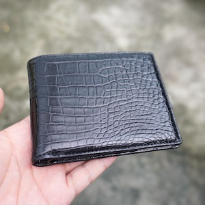 Op de afbeelding: Een zwarte bifold portemonnee met een gestructureerd krokodillenleerpatroon op het buitenoppervlak. De portemonnee heeft een rechthoekige vorm met gestikte randen en een glanzende afwerking, met een gedetailleerd schubbenpatroon op de voorzijde.