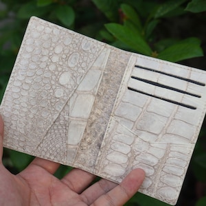 Può includere: Un portafoglio pieghevole realizzato in pelle testurizzata color bianco sporco con un distinto motivo a squame di rettile. L'interno presenta diverse fessure orizzontali per carte e una tasca verticale, mostrando un mix di texture a squame piccole e grandi.