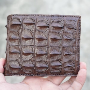 Peut inclure: Un portefeuille pliable marron foncé présentant un motif d'écailles proéminent, en relief et texturé sur toute la surface extérieure. Le portefeuille a une forme rectangulaire avec des coutures visibles sur les bords et une finition brillante.