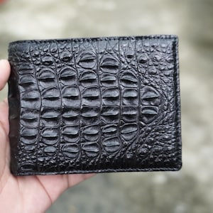 Puede incluir: Una cartera negra de dos pliegues con un patrón texturizado y elevado que imita la piel de cocodrilo. La superficie tiene un acabado brillante con escamas distintas y repetitivas que varían en tamaño a lo largo del exterior rectangular.