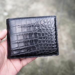 May include: A black bifold wallet featuring a glossy, textured crocodile skin pattern across the entire exterior surface. The wallet has a rectangular shape with stitched edges and a smooth, reflective finish that highlights the scale details.