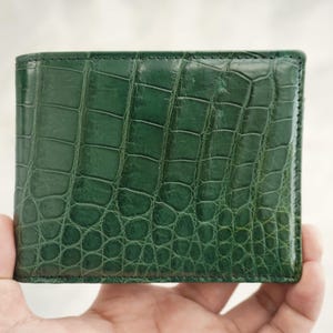 Puede incluir: Una cartera bifold de color verde bosque profundo con un acabado brillante y texturizado que imita la apariencia de la piel de cocodrilo. La cartera tiene costuras visibles a lo largo de los bordes y una forma rectangular.