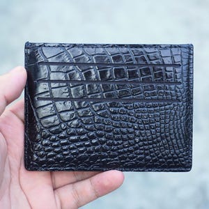 Può includere: Un porta carte rettangolare nero caratterizzato da una texture lucida in rilievo che imita la pelle di coccodrillo. La superficie frontale presenta tre fessure orizzontali per carte, con un motivo a scaglie uniforme su tutto l'esterno.