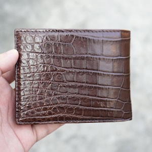 May include: A dark brown bifold wallet featuring a glossy, textured surface with a distinct embossed pattern that mimics the appearance of crocodile skin. The wallet has visible stitching along the edges and a rectangular shape with rounded corners.