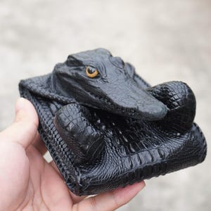 Può includere: Un portafoglio nero a due scomparti con una scultura tridimensionale di testa di coccodrillo sulla parte anteriore. Il portafoglio è realizzato con una trama simile a scaglie e presenta un dettaglio dell'occhio giallo ben visibile.