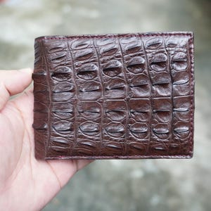 May include: A dark brown bifold wallet featuring a prominent, raised, textured scale pattern across the entire exterior surface. The wallet has a rectangular shape with stitched edges and a glossy finish that highlights the detailed, organic ridges.