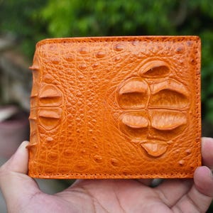 May include: A bright orange bifold wallet featuring a textured surface with prominent, raised, scale-like patterns. The exterior displays a distinct, repeating arrangement of large, rounded, and elongated bumps across the entire front panel.