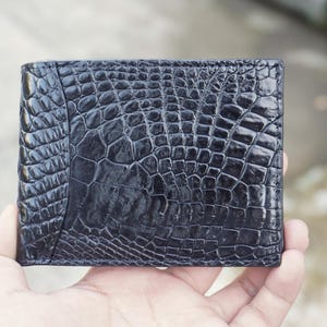 Könnte beinhalten: Ein dunkelblaues Bifold-Portemonnaie mit einer glänzenden, strukturierten Oberfläche und einem ausgeprägten Schuppenmuster, das an Reptilienhaut erinnert. Das Portemonnaie hat eine rechteckige Form mit sichtbaren Nähten an den Kanten.