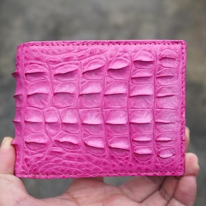 Könnte beinhalten: Ein leuchtend pinkfarbenes Bifold-Portemonnaie mit einem strukturierten, erhabenen Schuppenmuster auf der Außenfläche. Das Portemonnaie hat eine rechteckige Form mit sichtbaren Nähten an den Kanten und eine markante, feste Krokodilleder-Optik.