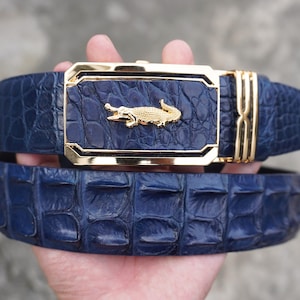 May include: A navy blue belt with a textured crocodile skin pattern. The belt features a rectangular gold-tone buckle with a matching navy blue inlay and a prominent gold-tone crocodile emblem centred on the buckle face.