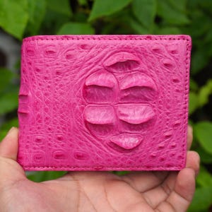 May include: A bright pink bifold wallet featuring a prominent, raised, textured pattern that mimics the appearance of crocodile or alligator skin, with a central section displaying larger, rounded, scale-like details across the front surface.