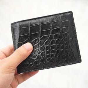 Könnte beinhalten: Ein schwarzes Portemonnaie im Bifold-Design mit einer glänzenden, strukturierten Oberfläche und einem ausgeprägten Prägemuster, das die Optik von Krokodilleder nachahmt. Das Portemonnaie ist rechteckig und hat sauber vernähte Kanten.
