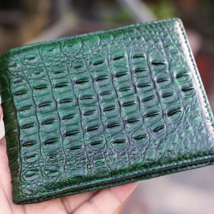 Può includere: Un portafoglio pieghevole di colore verde smeraldo intenso caratterizzato da una texture in rilievo che imita la pelle di coccodrillo. La superficie presenta una finitura lucida con dettagliati motivi a scaglie su tutto l'esterno e forma rettangolare.