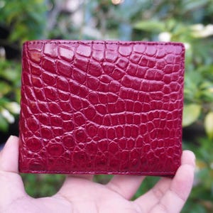 Può includere: Un portafoglio quadrato di colore rosso intenso e vivace. La superficie esterna presenta una distinta texture in pelle di coccodrillo, in rilievo e lucida, con un motivo a scaglie ripetuto sull'intero pannello frontale dell'accessorio in pelle.