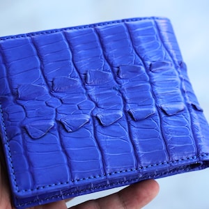May include: A vibrant royal blue bifold wallet featuring a prominent, raised, textured scale pattern across the exterior surface. The wallet is constructed with visible stitching along the edges and a distinct, segmented, reptilian-like skin texture.