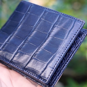 Puede incluir: Una cartera bifold de color azul oscuro con una superficie brillante y texturizada que presenta un patrón distintivo de escamas rectangulares. Los bordes de la cartera están cosidos con hilo azul oscuro a juego y acabado pulido.